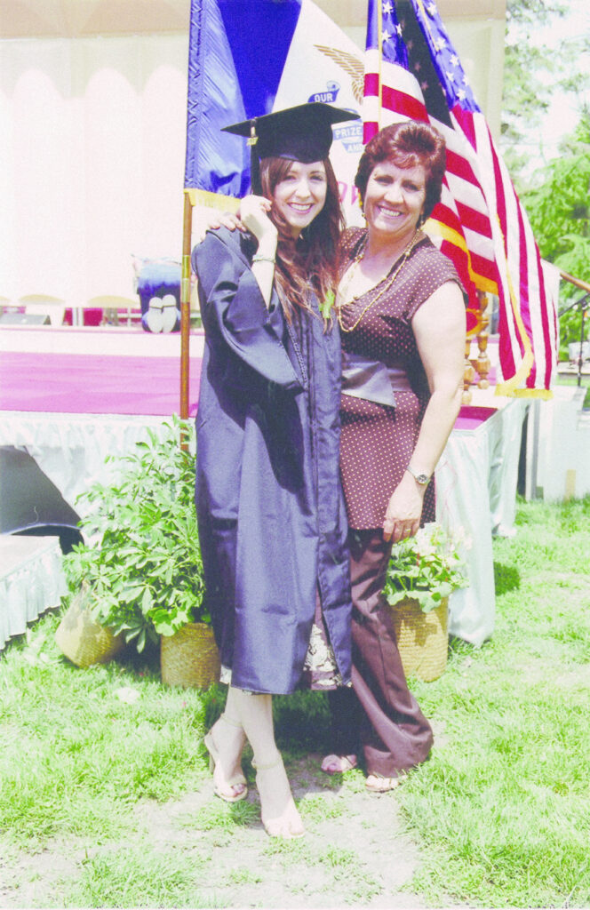 Photograph of two women, on the left a woman in her 20s with light skin and long straight brown hair. She is wearing a graduation cap and gown. Next to her a woman in her 40s wearing a brown top and pants, she has her arm around the younger woman. 