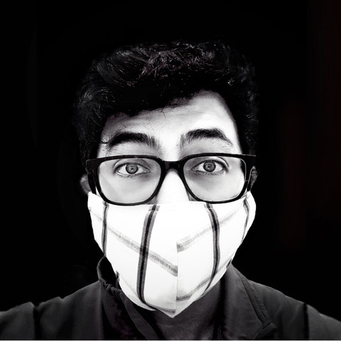 Black and white headshot of a man in his 30s who has heritage from Mexico with light skin tone and black short curly hair. He is wearing large black rimmed glasses, and a fabric mask with black and white lines.