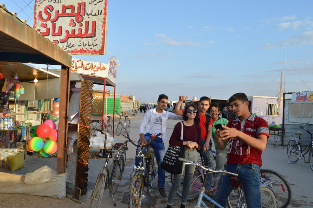 Photograph of a group of young people standing outside a store, most of them are sitting on bicycles. 