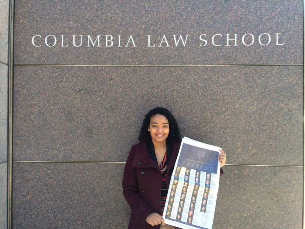 Photograph of a woman in her 20s who has medium skin tone and curly black hair, she is holding a New York Times and standing under a sign that says "Columbia Law School"