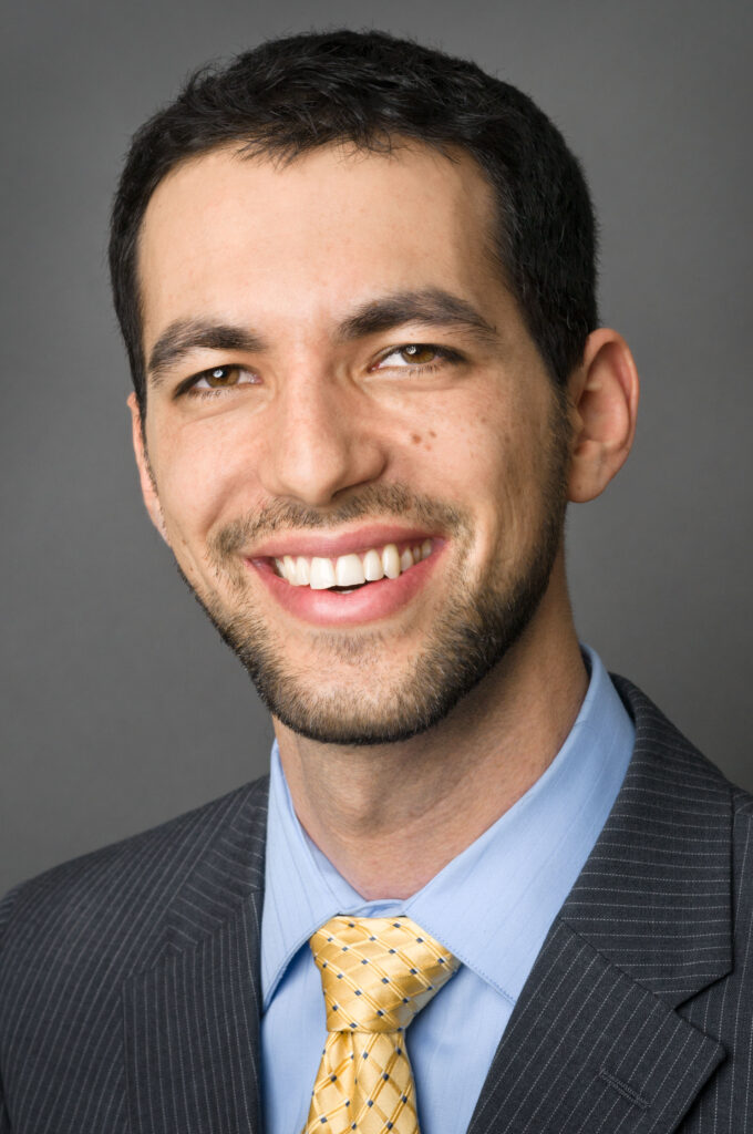 Headshot of a man in his 20s who has heritage from Israel and Romania with light skin tone, black short hair and a short-trimmed beard. He is wearing a pinstripe navy suit, light blue button down shirt and yellow tie. He is smiling at the camera.