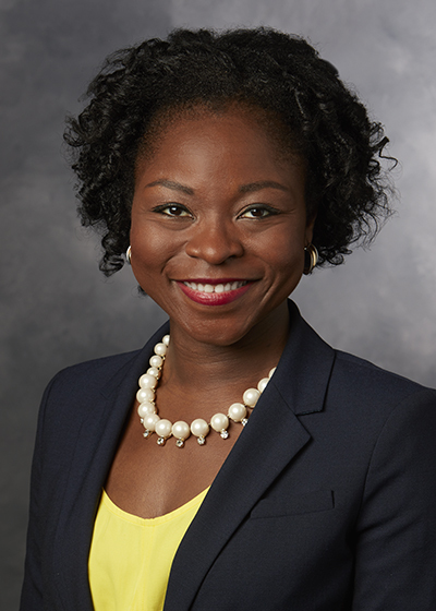 Headshot of a woman in her 30s who has heritage from Nigeria with dark skin tone and short bob coiled black hair. She is wearing a navy blazer, yellow blouse and large pearl necklace with gold earrings. She is smiling at the camera. 