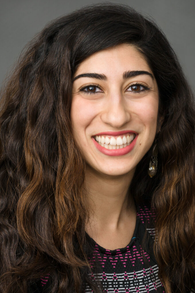 Headshot of a woman in her 20s who has heritage from Iran with light skin tone and long, wavy black with auburn highlights hair. She is wearing a black top with white, pink and teal shapes. She is smiling at the camera.