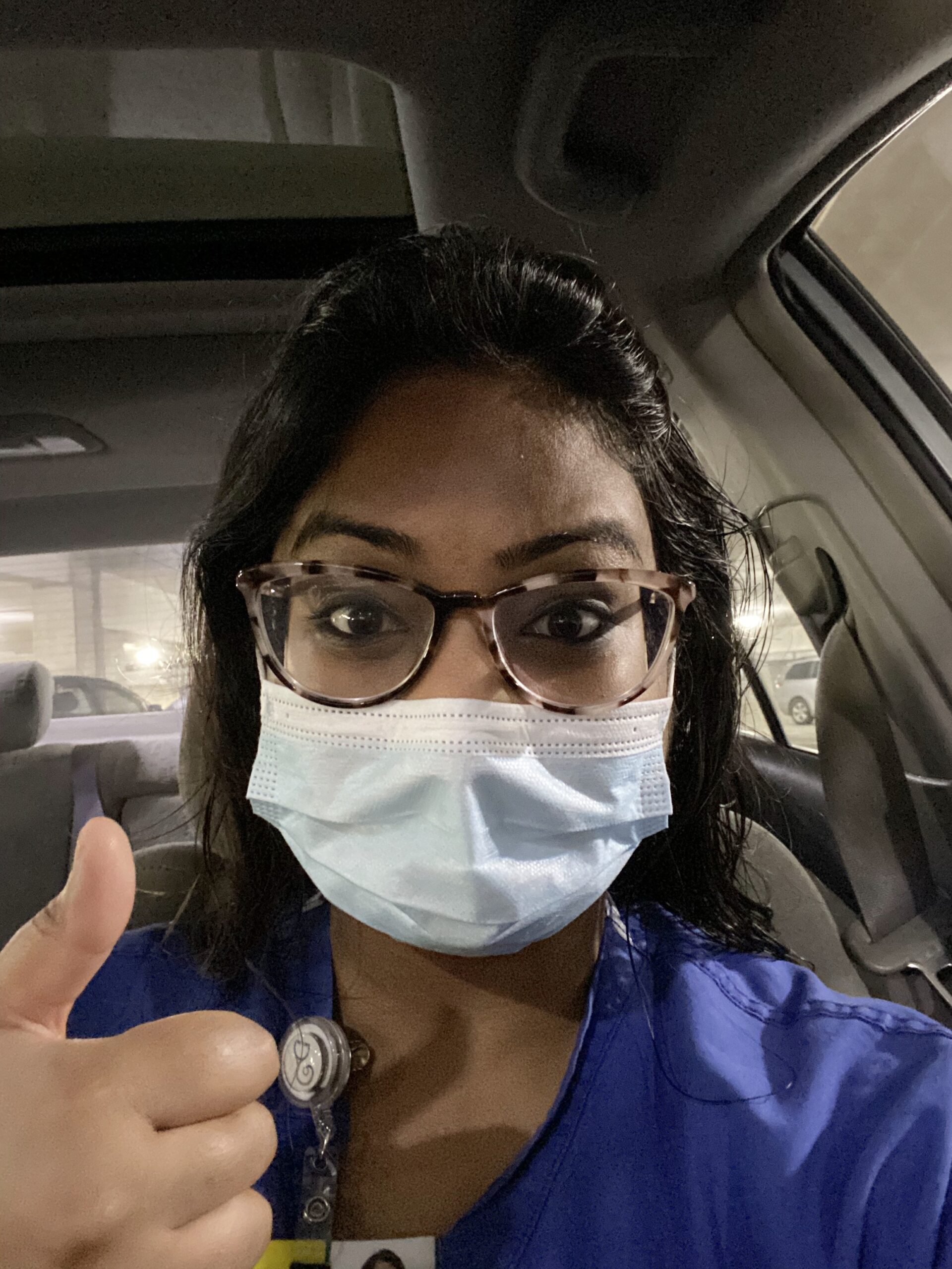 Selfie of a woman in her 30s who has heritage from India with dark-medium skin tone and black hair half pulled back. She is wearing blue scrubs, a surgical mask and large tortoiseshell glasses. She is looking at the camera and giving a thumbs up.