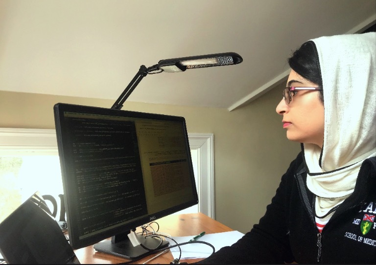 Photograph of a woman in her 20s, with heritage from Iran, she has light skin tone and only a swoop of black bangs can be seen peeking out from her white hijab.  She is wearing a black jacket. She looks at a computer screen away from camera.