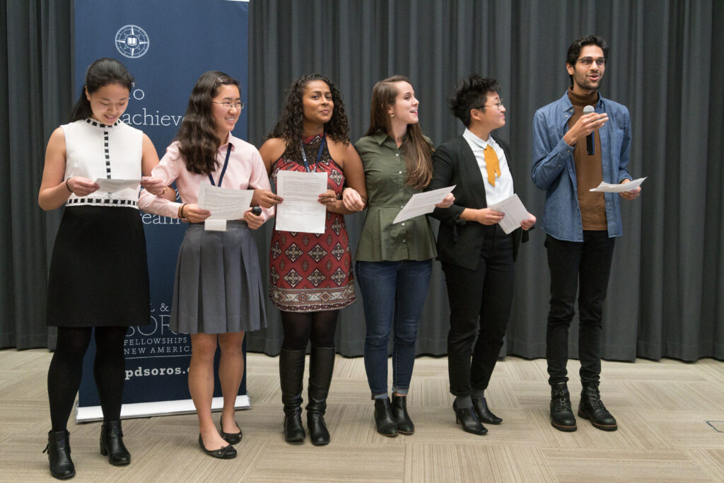 Photograph of four people standing arm in arm looking off camera right. They are all holding pieces of paper. 