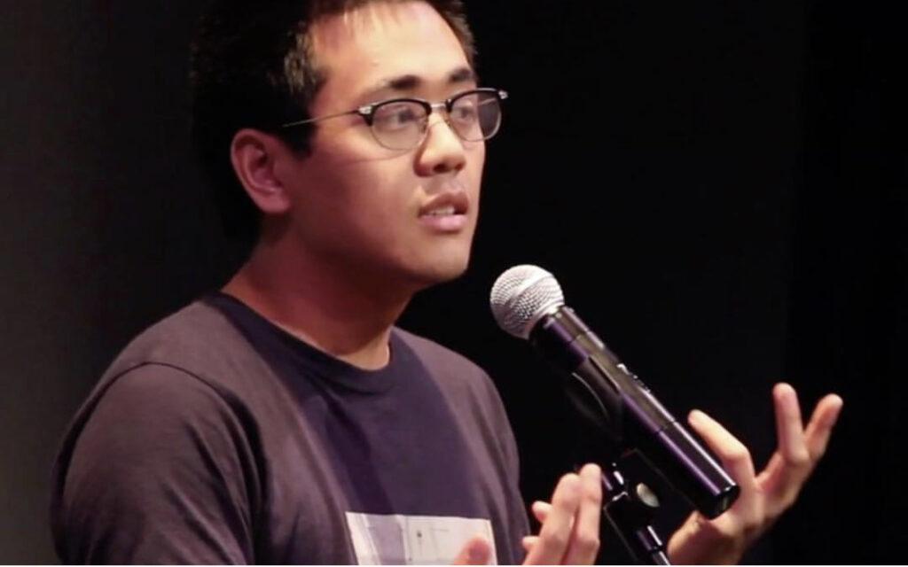 Photograph of a young man standing at a microphone, the background is black.