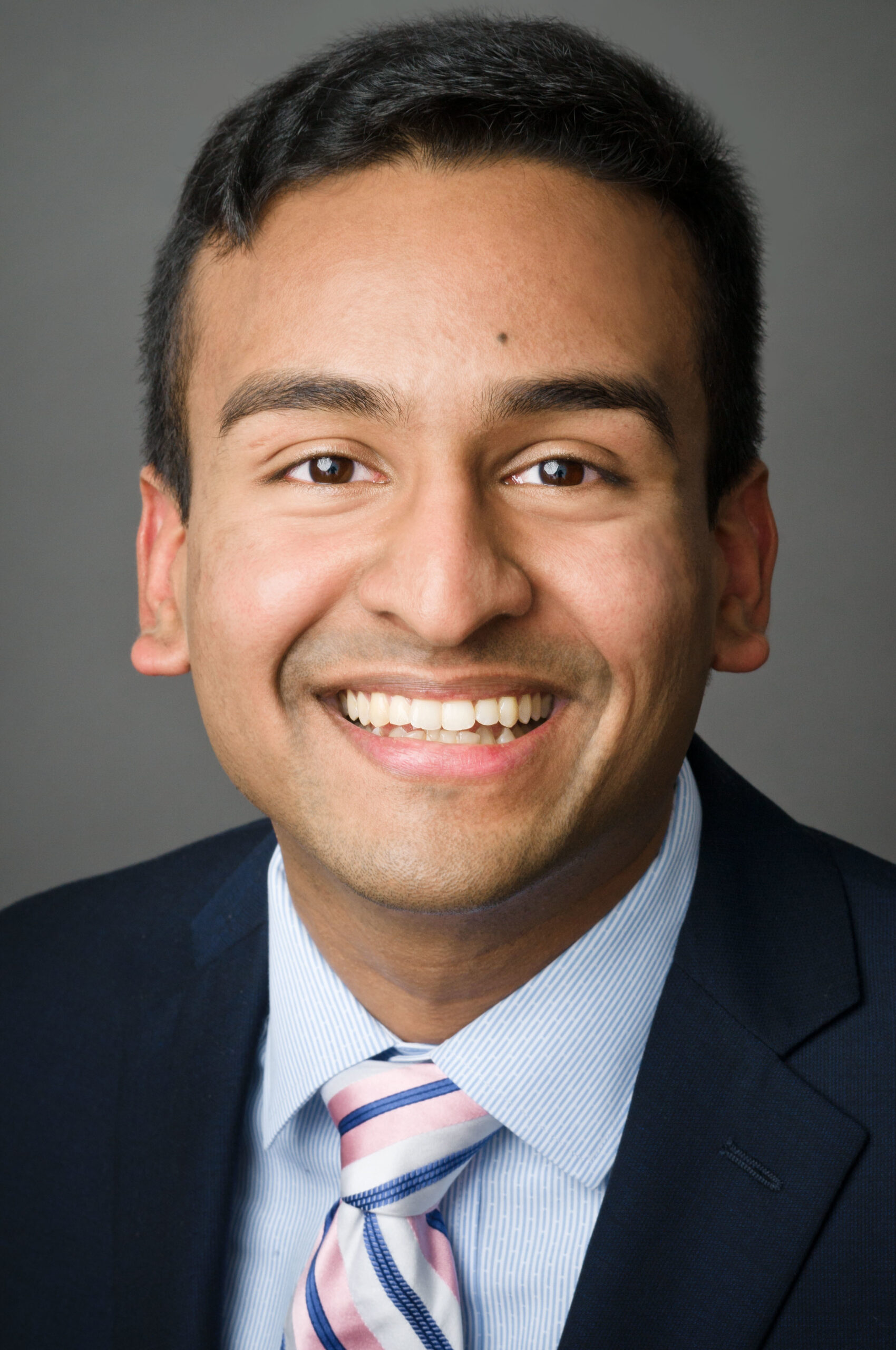 Headshot of a man in his 20s who has heritage from India, he has medium skin tone with short black hair. He is wearing a navy suit, blue and white stripped button up and pink, blue and white striped tie. He is smiling at the camera. 