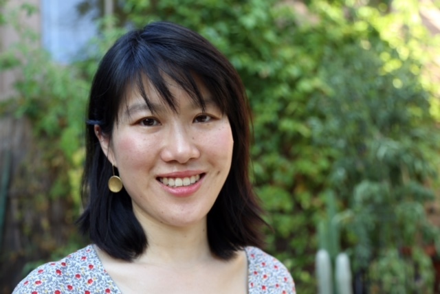 Headshot of a woman in her 30s who has heritage from Taiwan, she has light skin tone and shoulder length black hair with bangs. She wears a top with red and blue patterns, and brass dangly earrings. She is smiling at the camera. 