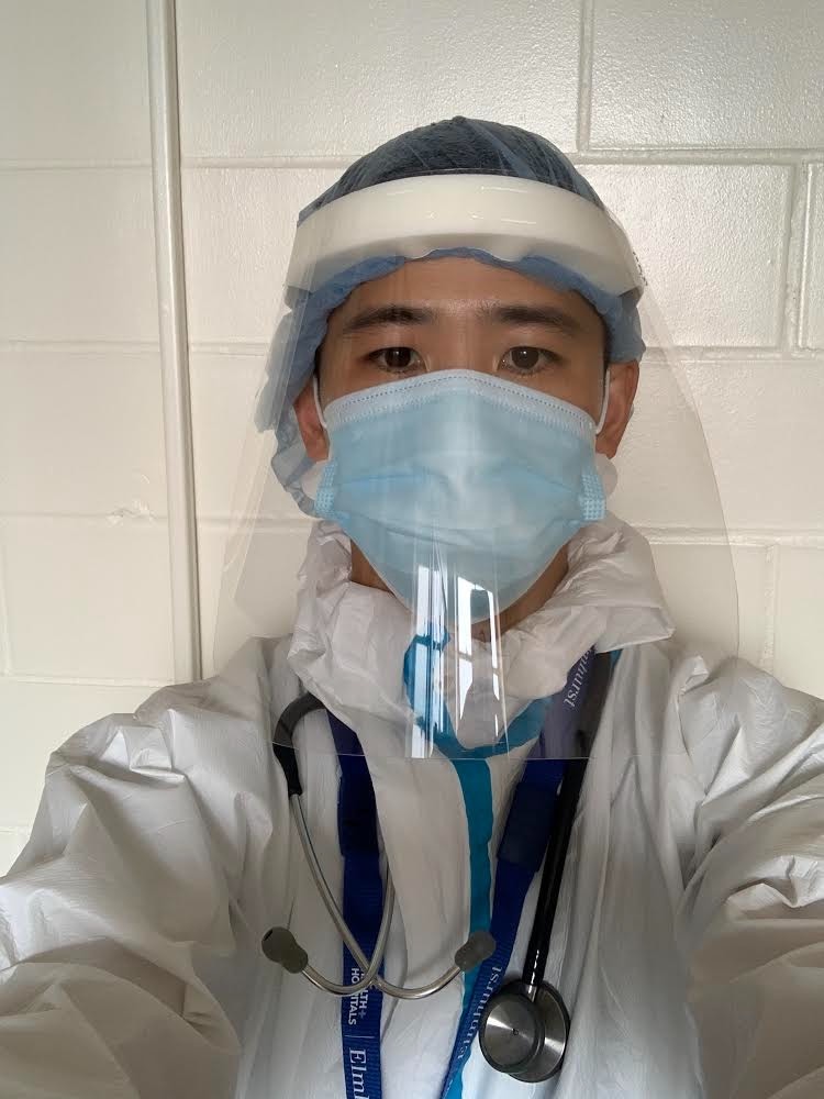 Selfie of a man in his 30s who has heritage from South Korea with light skin tone and black hair. He is dressed in full PPE (hairnet, face shield, surgical mask, outer layer on top of all clothing), he has a stethoscope and badge on a lanyard around his neck. 