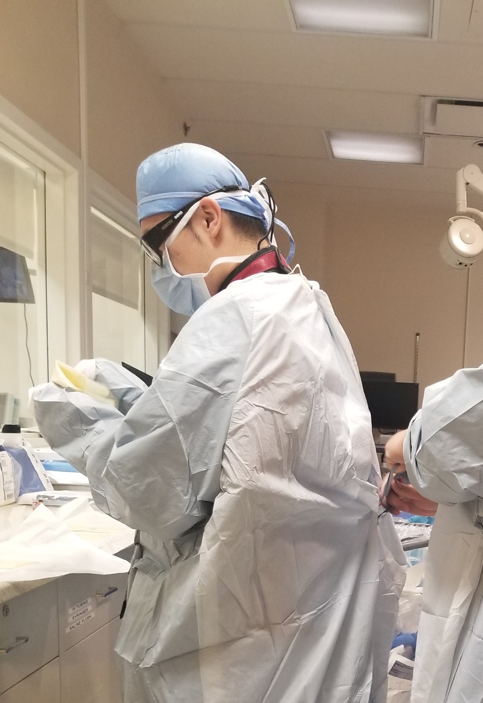 Photograph of a man in his 30s dressed fully in PPE (hair covering, surgical mask, glasses), we see his side and someone is tying the coverall closed, his hands are not seen as they are covered in PPE. They appear to be in a hospital.