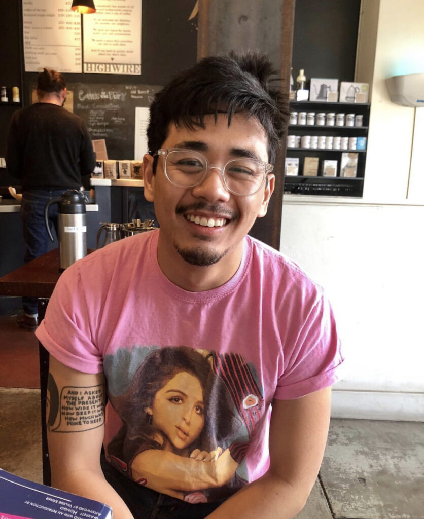 Photograph of a man in his 20s who has heritage from Cambodia with medium skin tone and black hair, slightly unkempt. He is wearing a pink t-shirt with a woman on it and large clear rimmed glasses. He is smiling at the camera. 