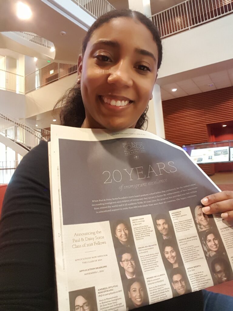 Photograph of a woman in her 20s with heritage from Dominican Republic with medium skin tone and tightly pulled back dark brown hair. She is holding a New York Times and smiling. 