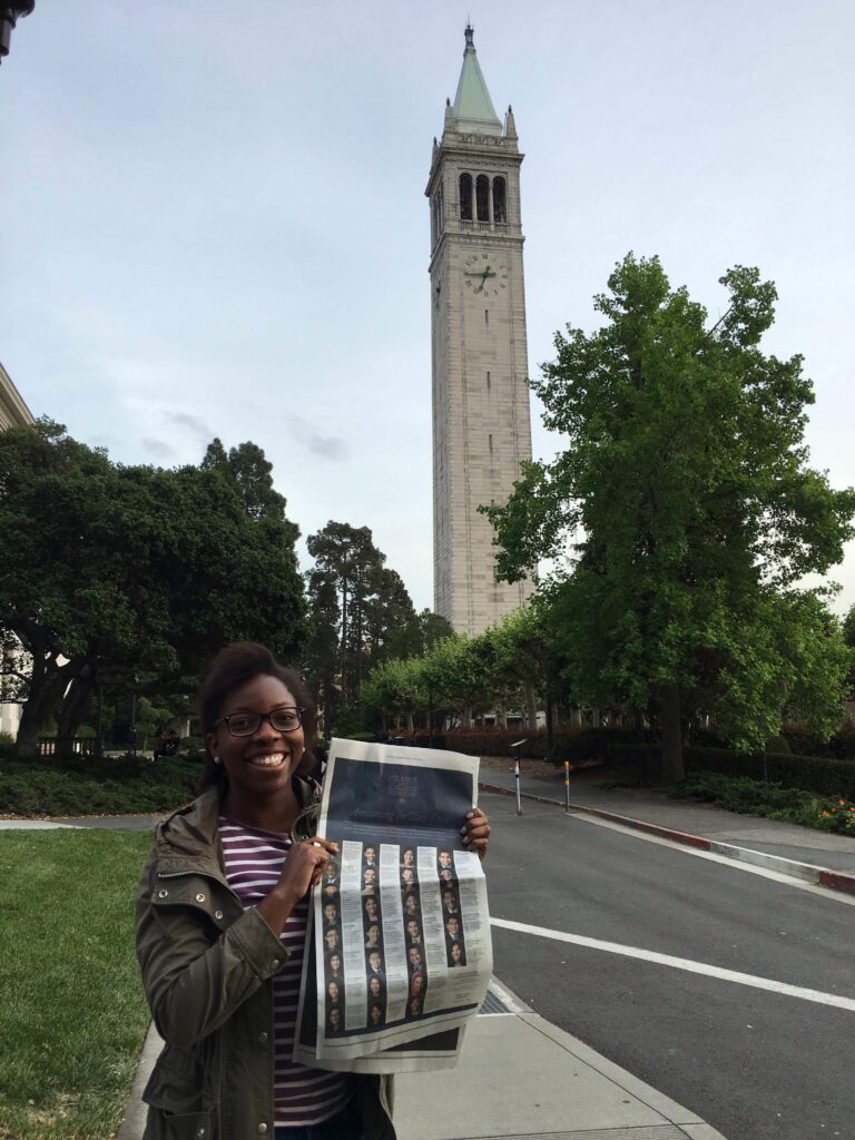 Photograph of a woman in her 20s with dark-medium skin tone and black hair, she is wearing a green jacket and white and red striped shirt, holding a New York Times. 