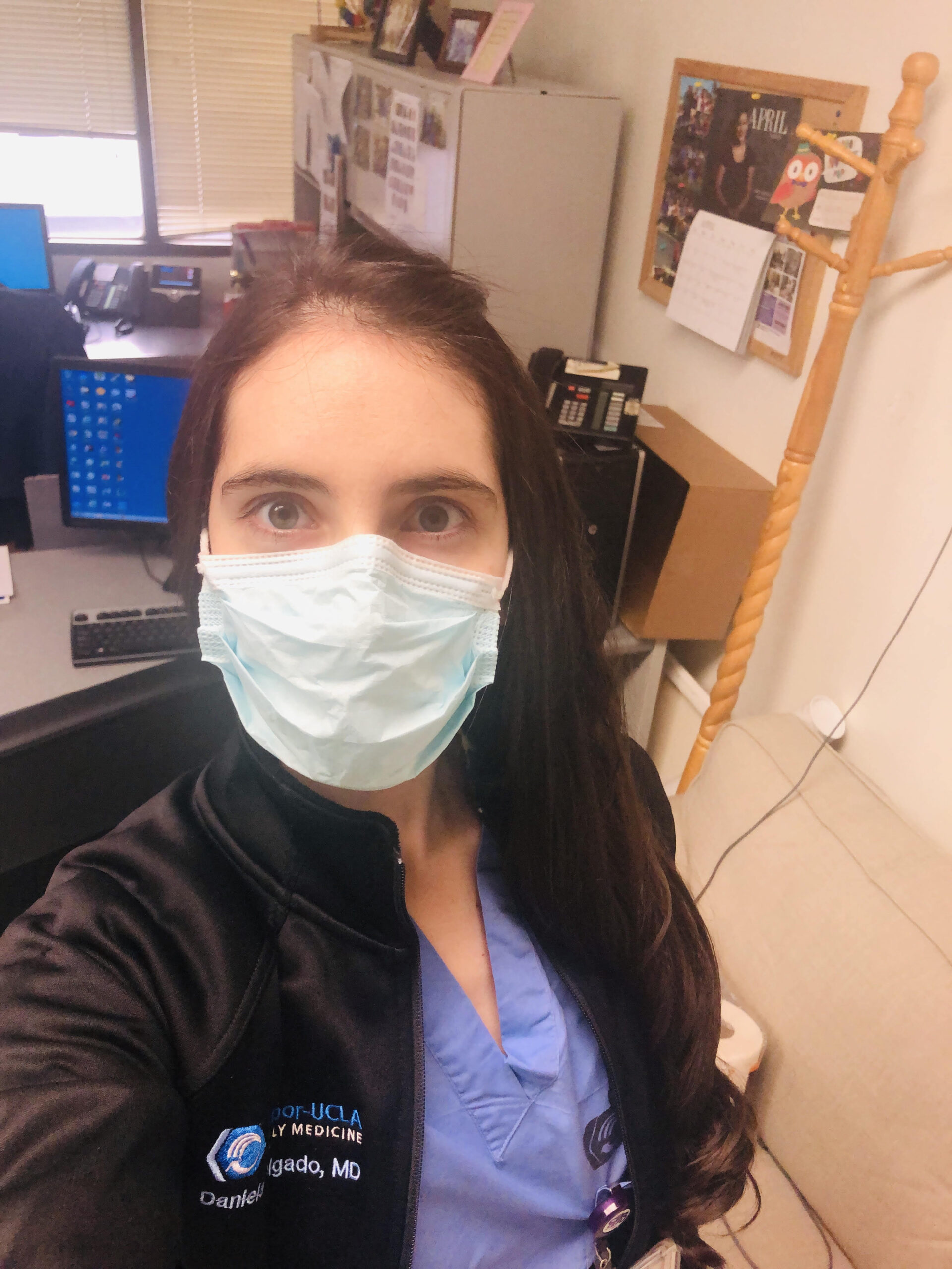 Selfie of a woman in her 20s who has heritage from Colombia, with light skin tone and long black straight hair pulled over her left shoulder. She is wearing blue scrubs with a black fleece jacket and a surgical mask, she is sitting in an office.