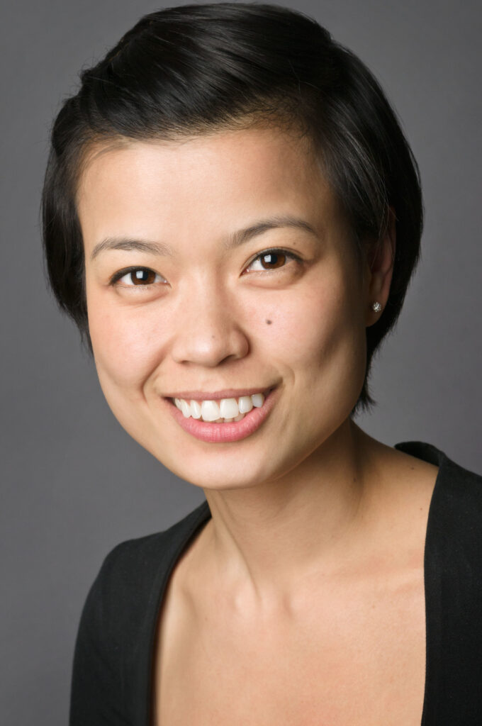 Headshot of a woman in her 20s who has heritage from Viet Nam with light skin tone and black pixie length hair slightly pulled back. She is wearing a black scoop neck top with diamond stud earrings and smiling at the camera. 