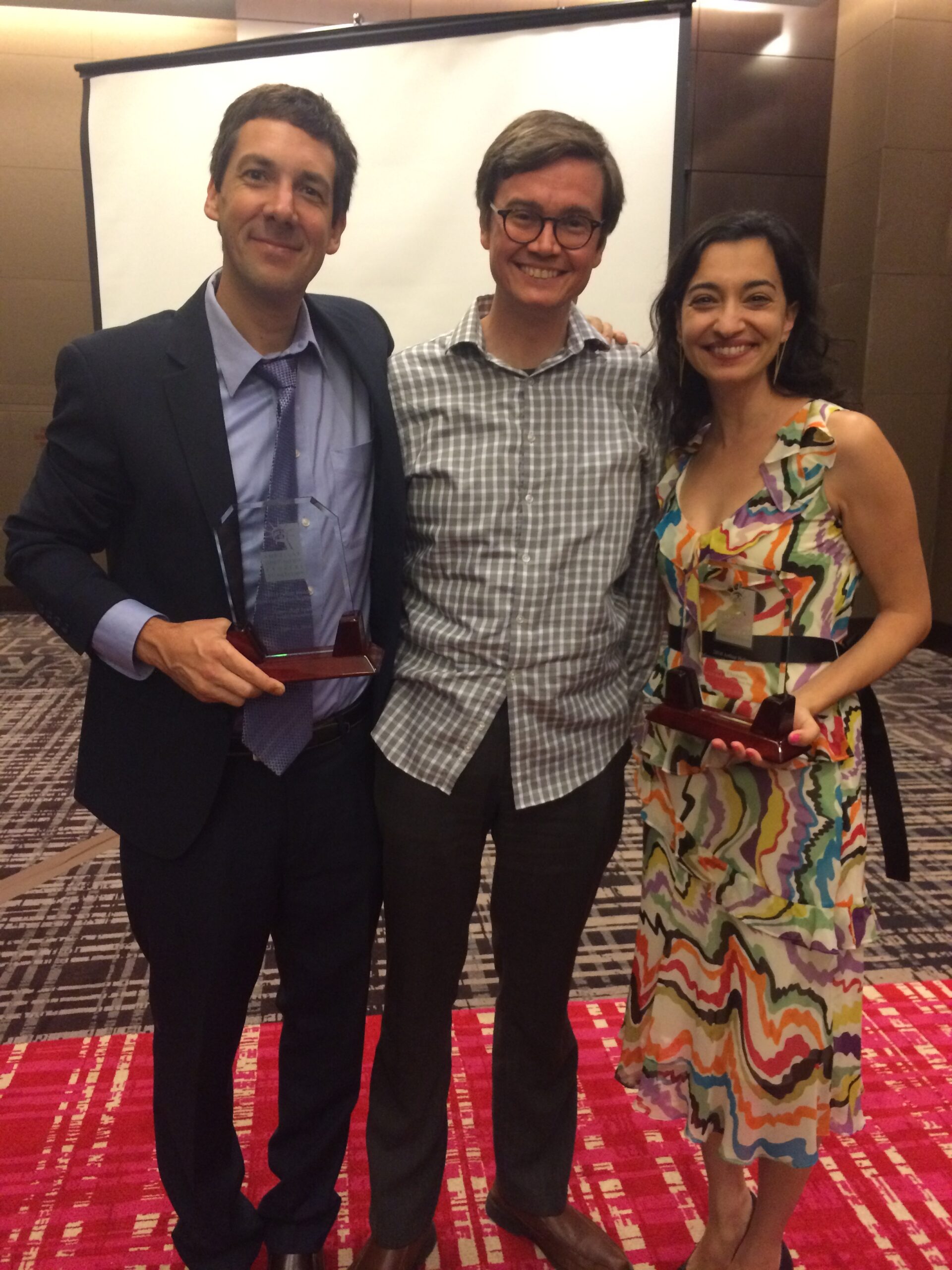 Photograph of three people standing in what appears to be a hotel ballroom, the man on the left and the woman on the right are holding awards that have a wood base and glass top. 