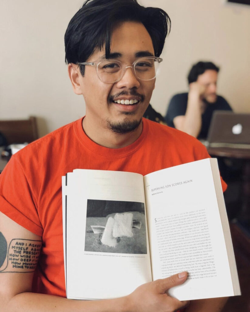 Photograph of a man in his 20s who has heritage from Cambodia with medium skin tone and longer black hair. He is wearing a bright red t-shirt and large clear framed glasses. He is holding a book open and smiling at the camera. 