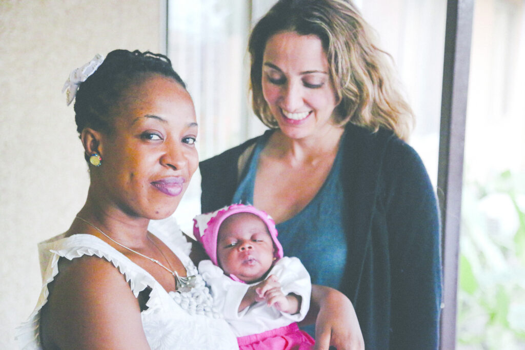 Photograph of two women, in the foreground a woman with medium-dark skin tone and black hair pulled back, she is wearing a white blouse and holding a small baby dressed in pink and white, she is smiling at the camera. The woman in the back has light skin tone and blonde hair, wearing a teal top and navy sweater, she is looking at the baby and smiling. 