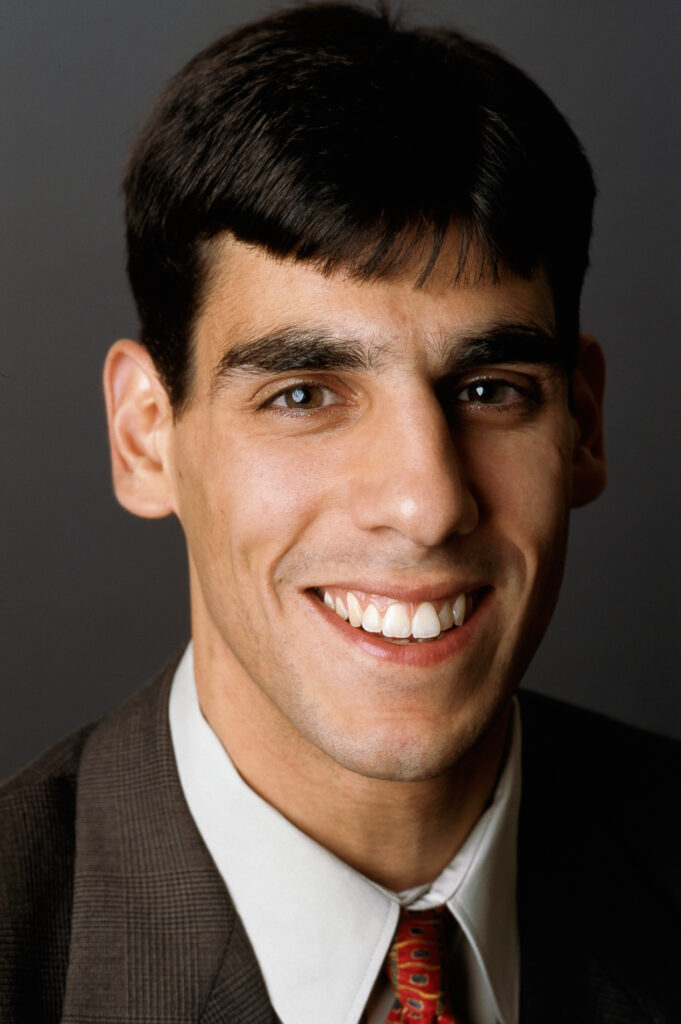 Headshot of a man in his 30s with heritage from Lebanon he has light-medium skin tone and black short cut hair. He is wearing a brown suit, white button up and red tie. He is smiling at the camera.