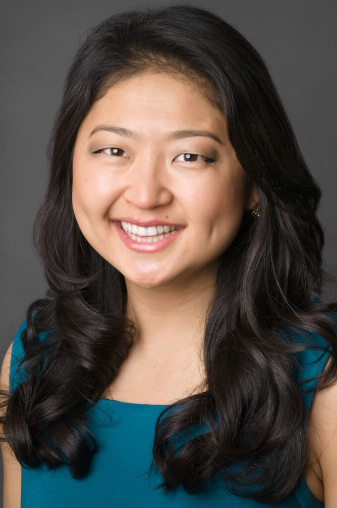 Headshot of a woman in her 20s who has heritage from Uzbekistan with light skin tone and passed shoulder length curled black hair. She is wearing a dark teal sleeveless blouse and gold earrings and smiling at the camera.