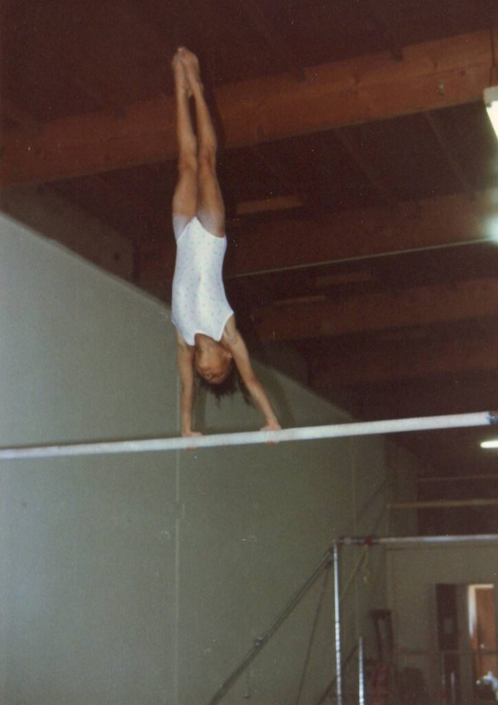 Old photograph of a young girl holding herself upright on a bar, she is wearing a white leotard. 