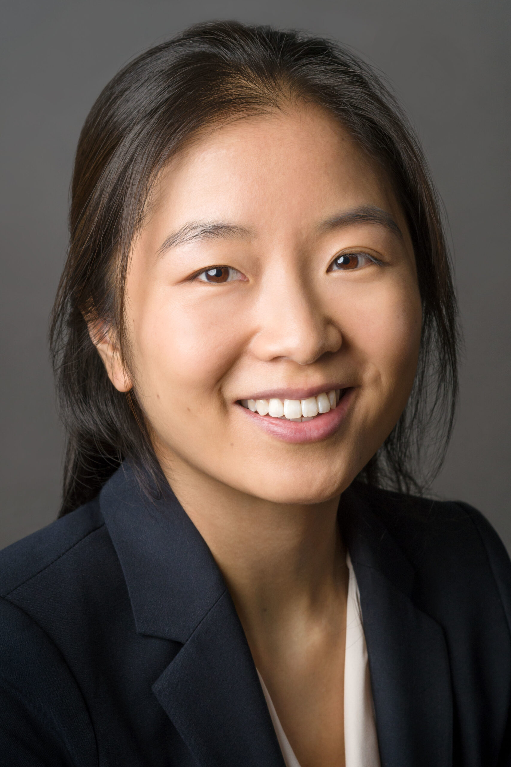 Headshot of a woman in her 20s who has heritage from China. She has light-medium skin tone with long dark brown hair pulled back and loosely falling toward her face. She is wearing a navy blazer and white blouse. She is smiling at the camera.