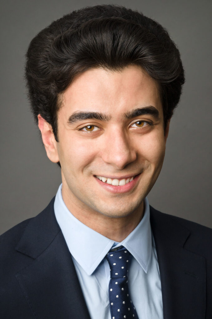 Headshot of a man in his 20s who has heritage from Iran with light skin tone and black combed back hair. He is wearing a navy suit, light blue button up shirt and a navy tie. He is smiling at the camera.