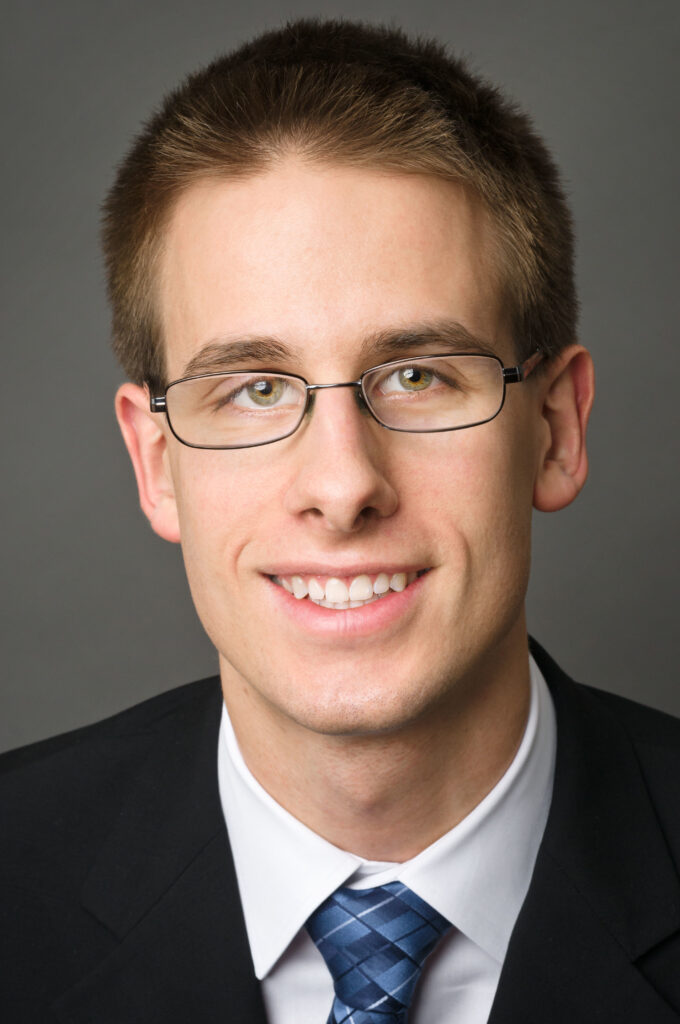 Headshot of a man in his 20s who has heritage from Poland with light skin tone and crew cut blonde hair. He is wearing a black suit, white button up shirt, blue tie and has thin rimmed oval glasses. He is smiling at the camera.