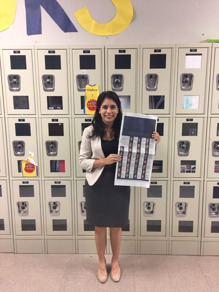 Photograph of a woman in her 20s who has heritage from Mexico with light skin tone and black hair, she is wearing a black dress and white blazer. She is standing in front of lockers and holding a New York Times.