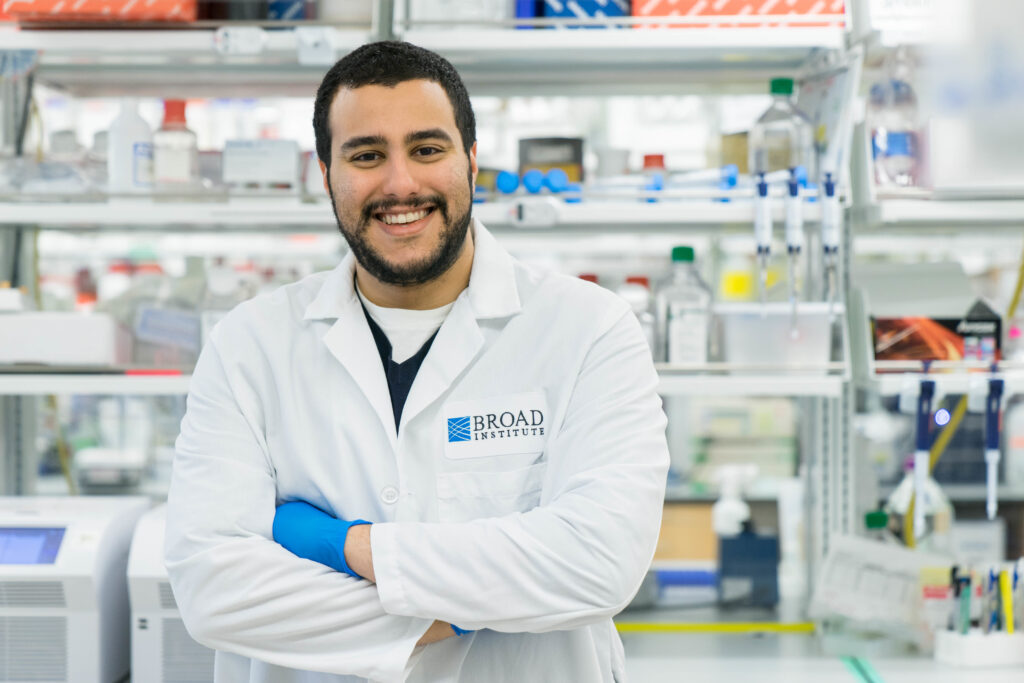 Half body photograph of a man in his 20s with heritage from Jordan and Kuwait he has light skin tone, black short hair and a short beard. He is wearing a white lab coat and blue gloves. He is standing in a lab with his arms crossed over his chest and smiling at the camera. 