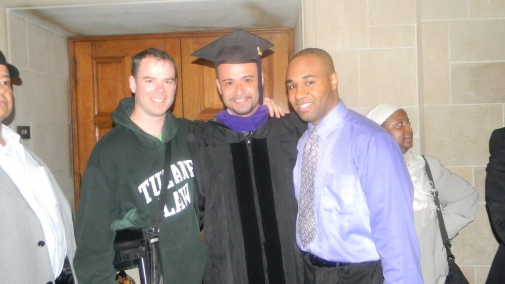 Photograph of three men in their 20s with their arms around each other. The man in the middle is wearing a black graduation gown and cap with a purple hood.