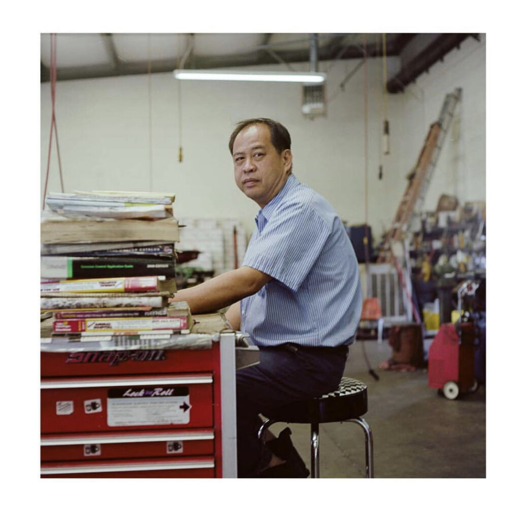 Old photograph of a man in his 40s wearing navy pants and a light blue short sleeve button up shirt. He is sitting at a work bench that is stacked high with books. A garage / workspace can be seen behind him.