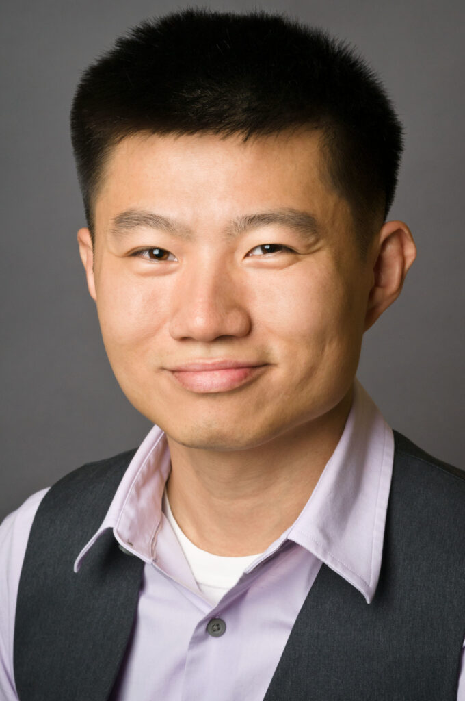 Headshot of a man in his 20s who has heritage from China with light-medium skin tone and black short hair. He is wearing a light purple button up and a grey vest. He is smiling at the camera. 