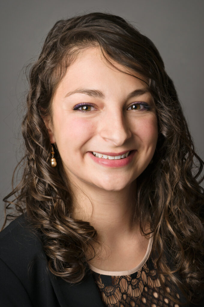 Headshot of a woman in her 20s who has heritage from Mexico with light skin tone and curly, light brown shoulder length hair. She is wearing a black blazer and black lace top with dangly pearl earrings. She is smiling at the camera. 