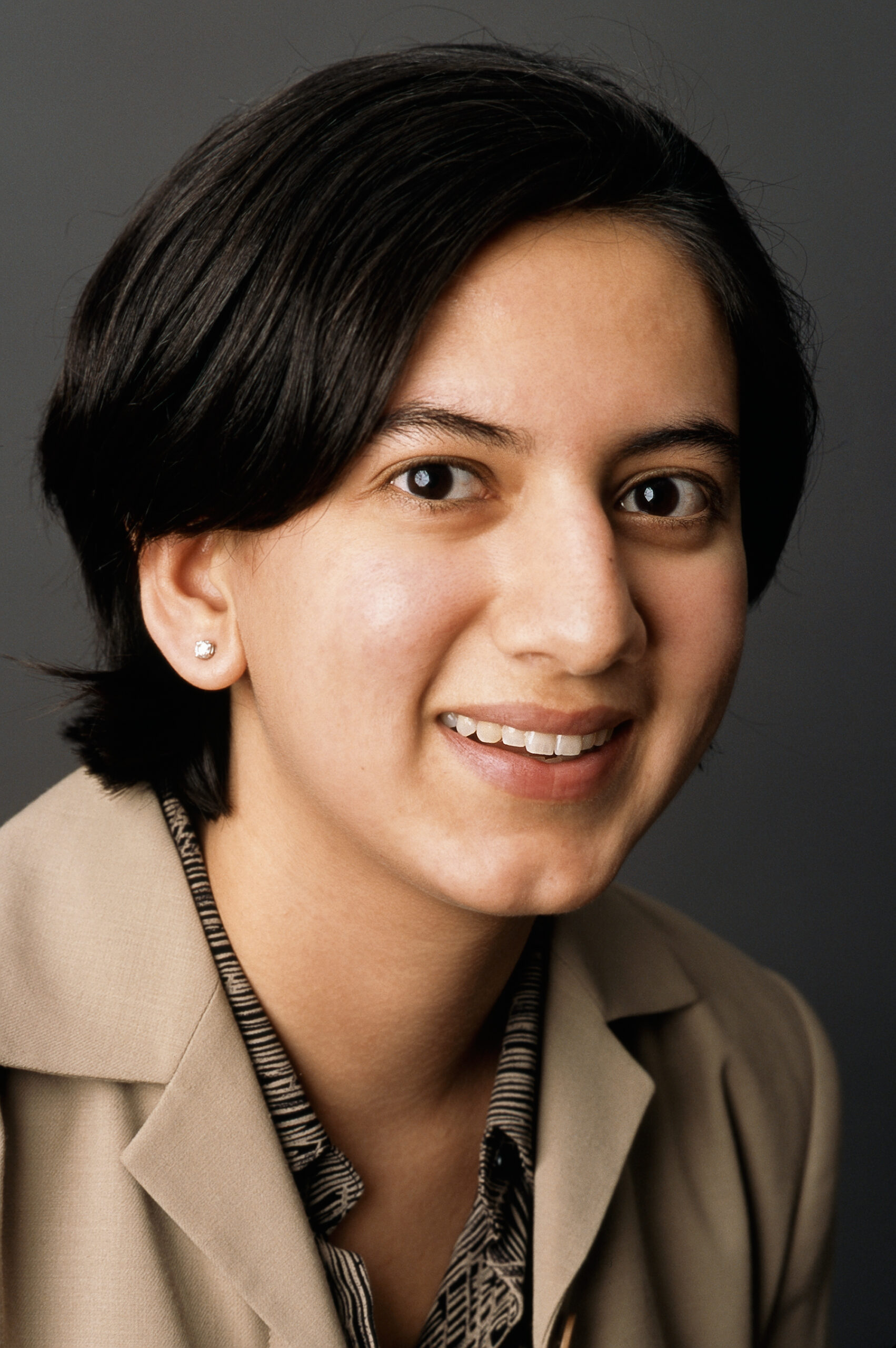 Headshot of a woman in her 20s who has heritage from India, she has light skin tone and black medium length hair. She is wearing a tan blazer with a tan and black button up, diamond stud earrings and is smiling at the camera.