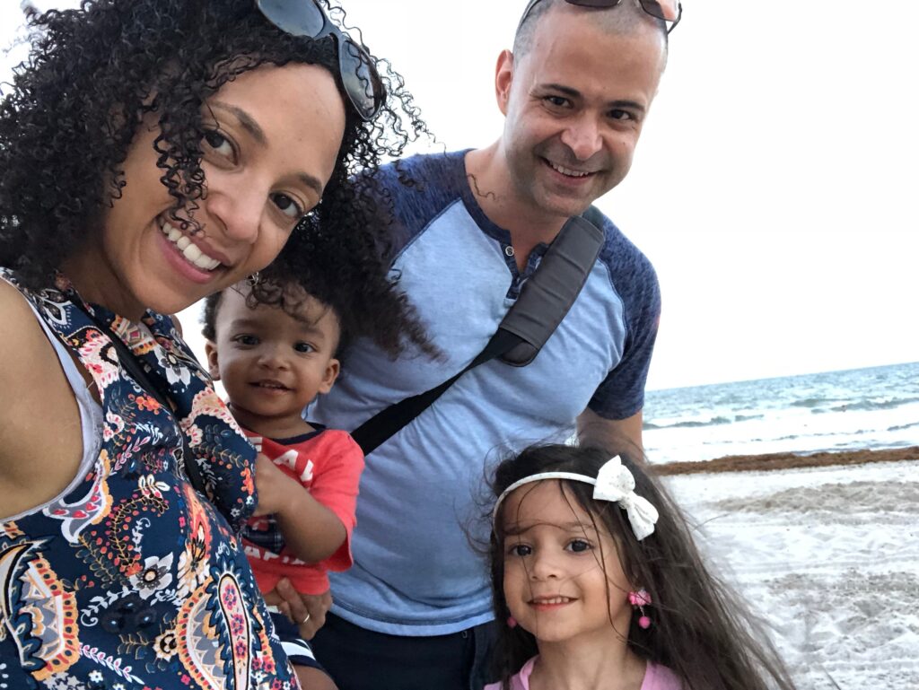 Selfie of a family, mom, dad and two young kids. The beach can be seen behind them. 