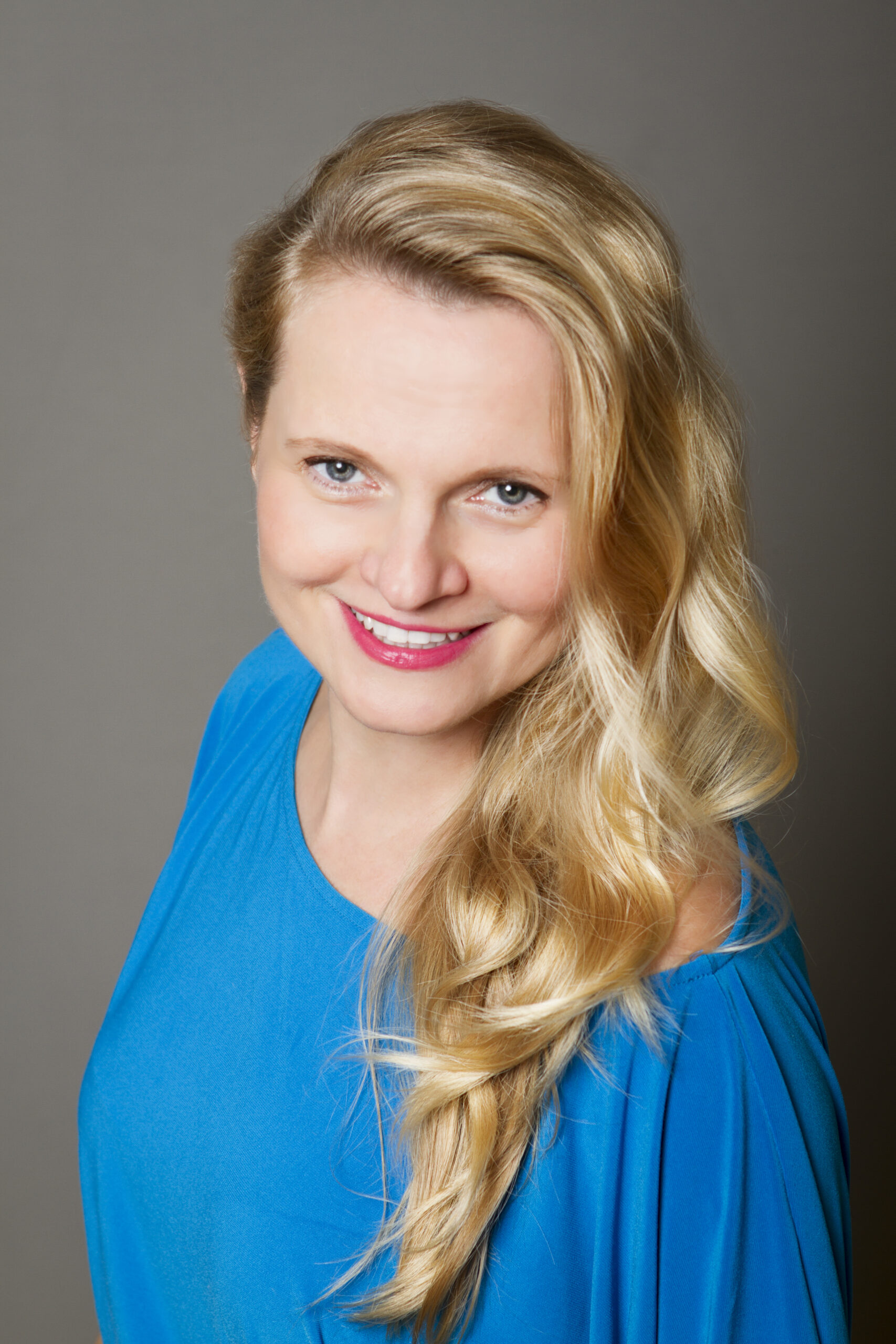 Headshot of a woman in her 30s with heritage from Poland she has light skin tone and wavy, long blonde hair. She is wearing a bright blue boat neck dress. She is looking slightly up at the camera and smiling.