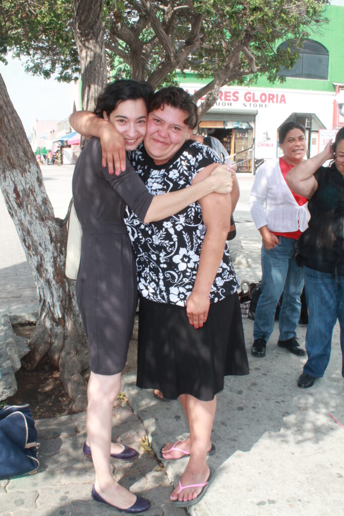 Photograph of two women, on the right a woman with brown hair and light skin tone wearing a black and white patterned top, black skirt and pink flip flops. The woman on the left who has heritage from Iran has light skin tone and short black curly hair. She is wearing a grey dress. The women are tightly embracing and looking at the camera. 