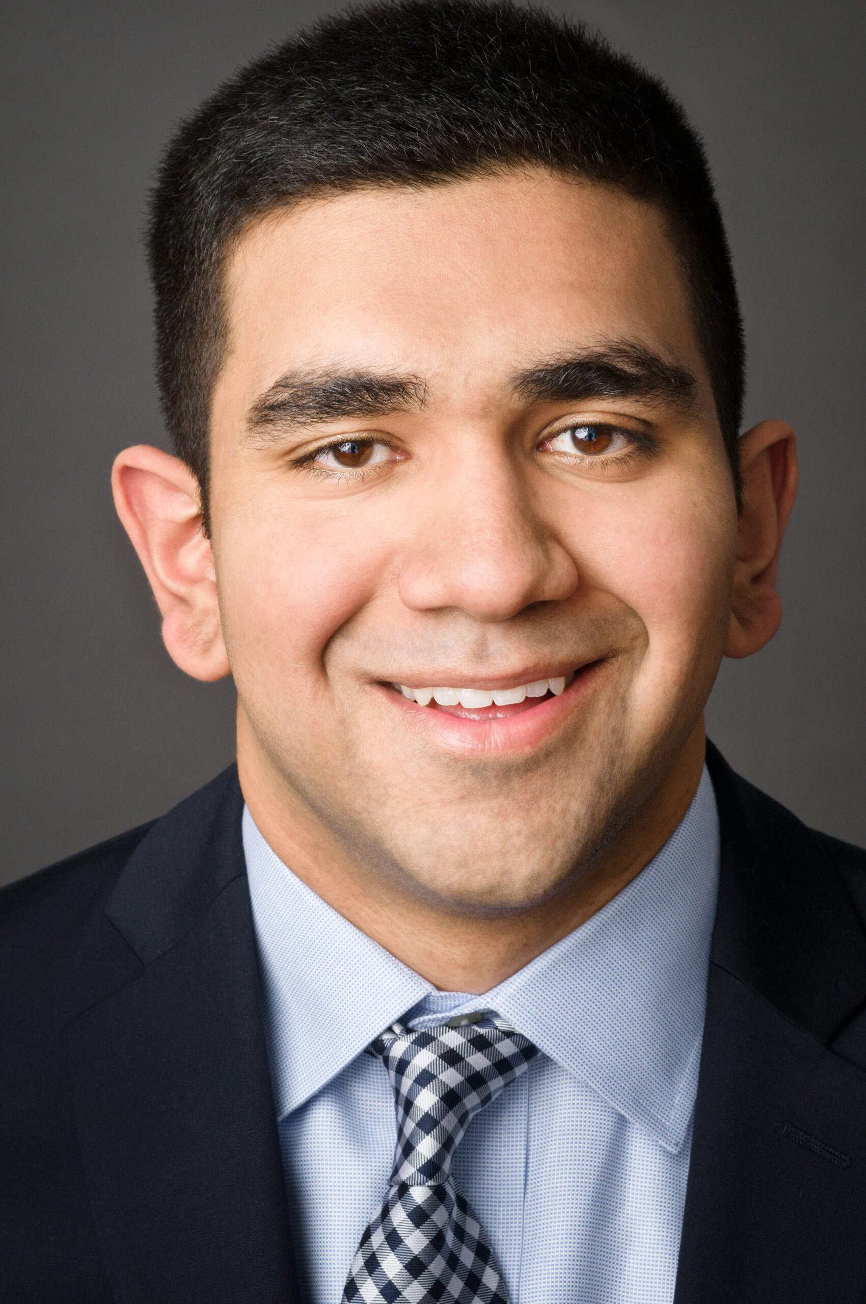 Headshot of a man in his 20s who has heritage from India with light skin tone and buzzed black hair. He is wearing a black suit, light blue button up shirt and blue and white gingham tie. He is smiling at the camera.