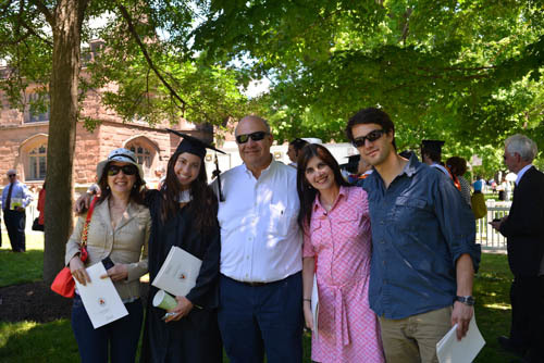 Photograph of five people standing under a tree on a lawn. Second to the left is a woman in her 20s wearing a graduation gown and cap. 