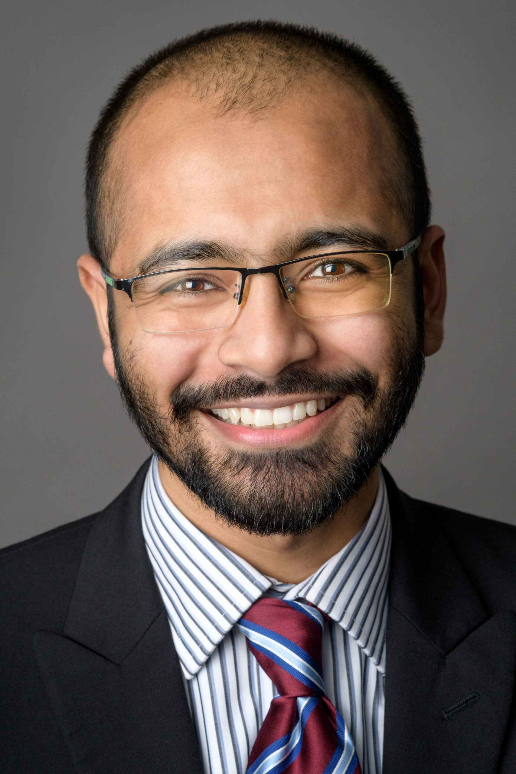 Headshot of a man in his 20s who has heritage from Pakistan, who has medium skin tone, black buzzed hair, and a short beard. He is wearing a black suit, blue and white striped shirt, red and blue tie and frameless rectangle glasses. He is smiling at the camera.