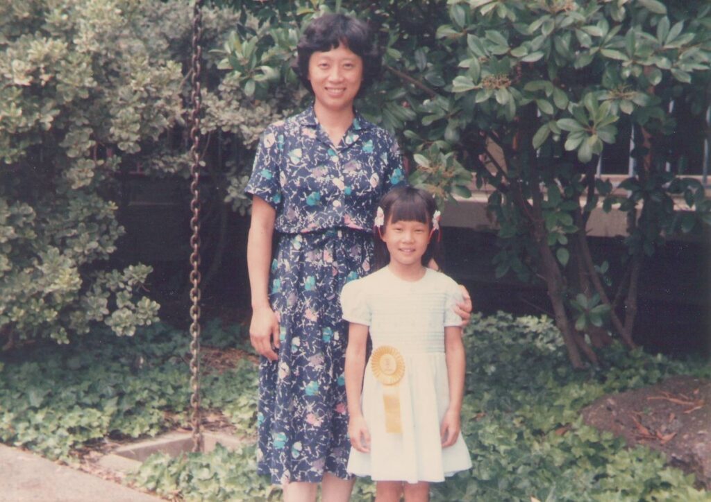 Old photograph of a woman and a young girl. The woman is wearing a blue floral dress and the young girl had a light blue dress and a yellow ribbon pined to her waist. 