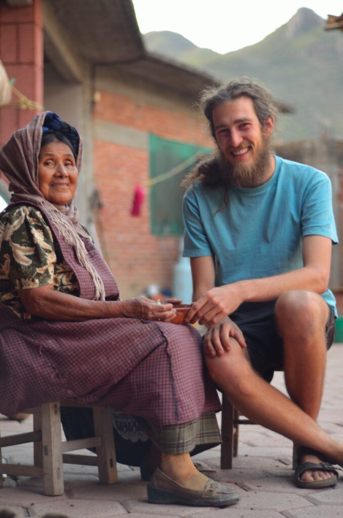 Photograph of a woman with medium skin tone and black hair pulled back and under a scarf, sitting down with a bowl in her lap, she is smiling at the camera. Next to her a man in his 20s with light skin tone, long light brown hair and a long beard sits smiling at the camera. 