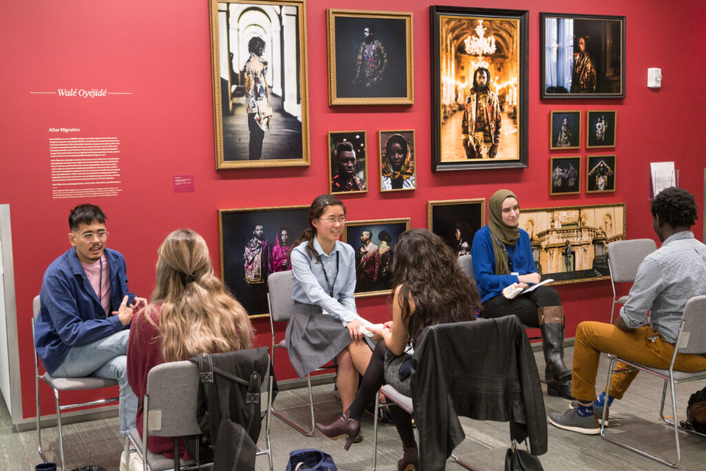 Photograph of six people sitting in chairs, facing one another making three groups of two. They appear to be in discussion. They are sitting in a room with a red wall and gallery style photos on the wall. 