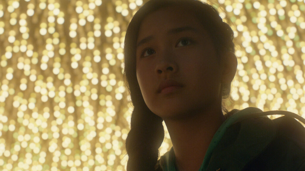 Movie still of a young woman with a single braid in her hair over her shoulder, looking past the camera; the background is gold lights. 