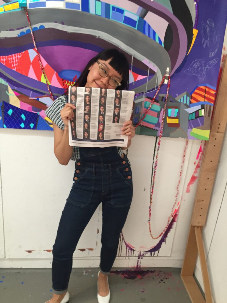 Photograph of a woman in her 20s with light skin tone and black hair with bangs, she is wearing overalls and a white and green gingham shirt, holding a New York Times. She is standing in front of a very colorful painting.