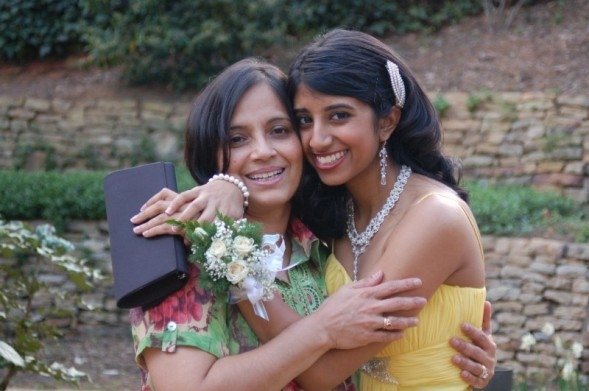 Photograph of two women, the woman on the right is in her 20s who has heritage from India, with medium skin tone and curled black hair, she is wearing a yellow dress with a large bejeweled necklace and dangly earrings. She is hugging a woman in middle-age with light skin tone and black hair, wearing a green and red top.