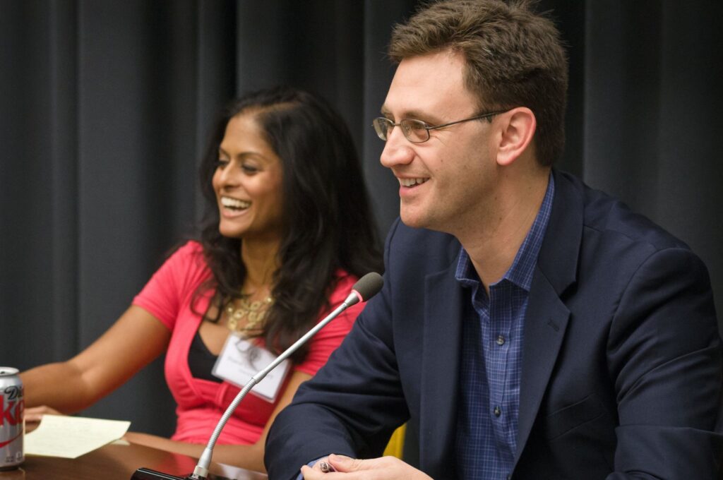 Photograph of a man in his 30s who has heritage from Czech Republic with light skin tone and short brown hair. He is wearing a navy blazer and blue button up shirt. He is seated at a microphone and looking off camera. A woman in her 30s sits next to him, laughing.