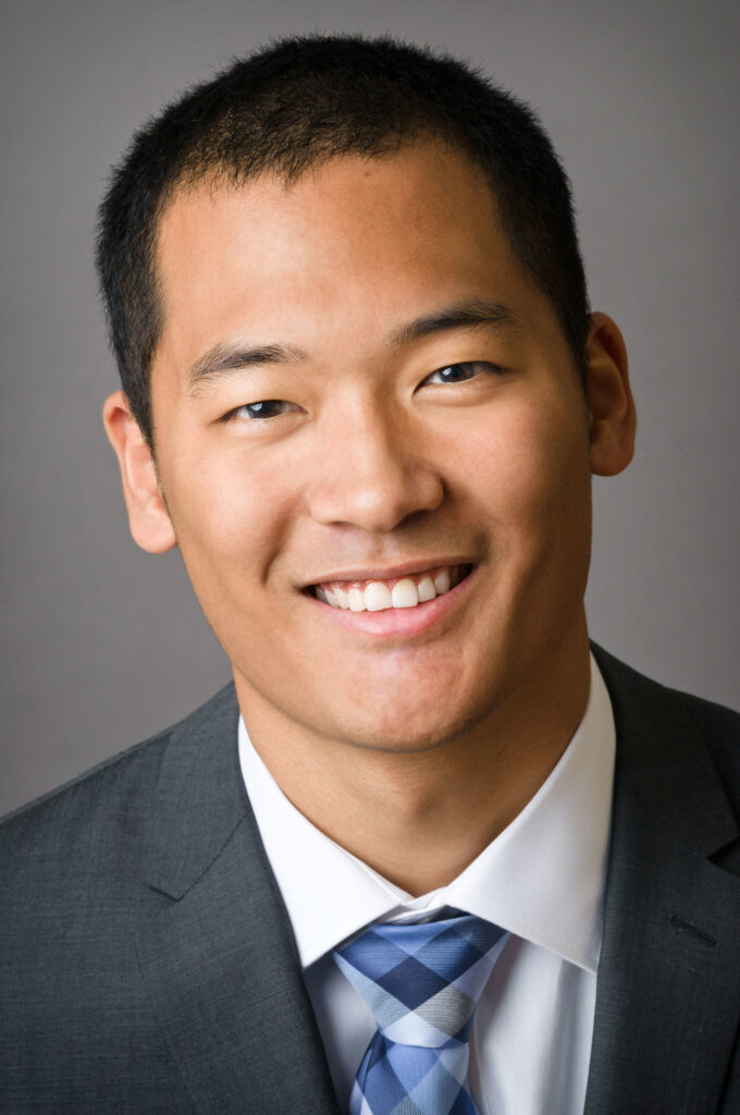 Headshot of a man in his 20s who has heritage from China with light-medium skin tone and buzzed black hair. He is wearing a grey suit, white button up shirt and blue tie.